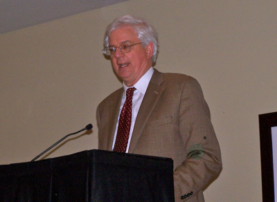 An adult wearing a tan jacket speaks behind a lectern.