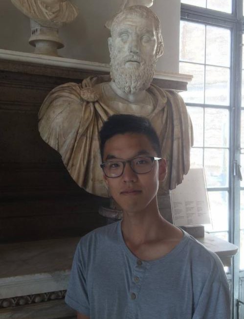 An adult stands in a museum beside a large sculpted bust.