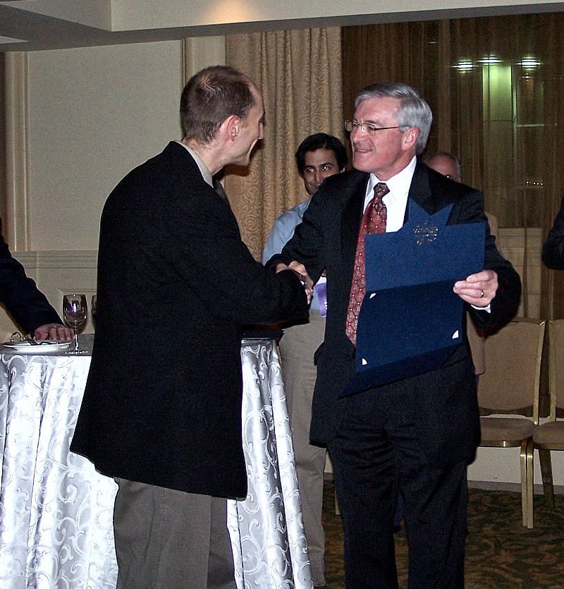 Two adults converse indoors, one holding an open folder.