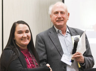 Two adults wearing conference badges stand together, one holding a microphone.