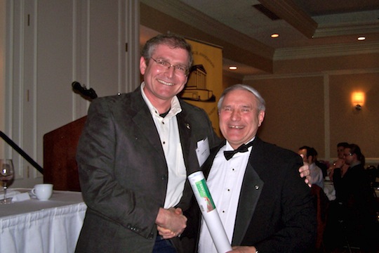 Two adults in formal dress stand together, one holding a rolled document.