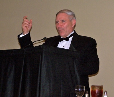 An adult in formal dress gestures while speaking at a microphone.