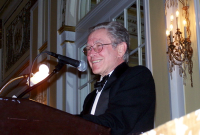An adult in formal dress smiles behind a lectern and microphone.
