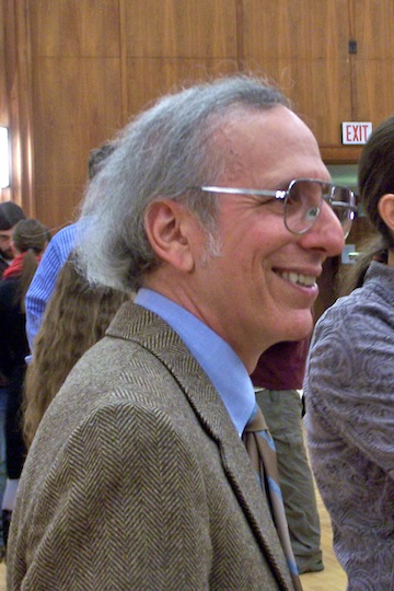 An adult with glasses and long gray hair smiles at an indoor gathering.