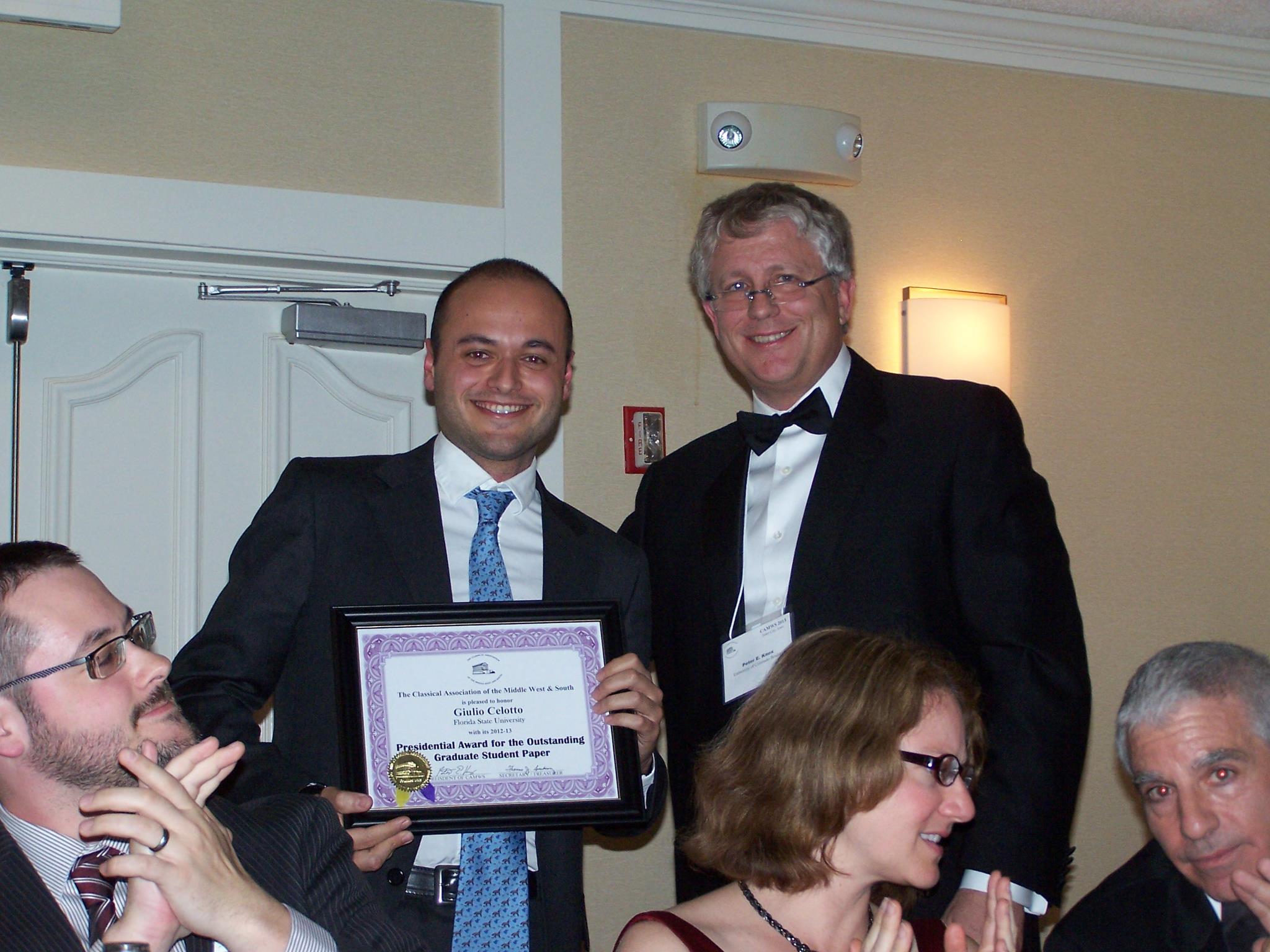 Two standing adults pose with a framed certificate beside seated banquet guests.