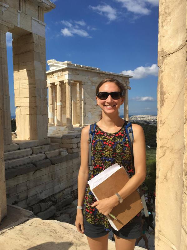 An adult holding a notebook stands beside stone columns at an ancient site.