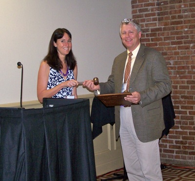 Two adults exchange a gavel beside a lectern.